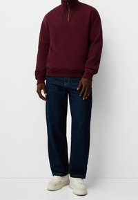 Sudadera burdeos con media cremallera y cuello acanalado, combinada con jeans azul oscuro de pierna recta y zapatillas cremas. Estilo casual y cómodo.