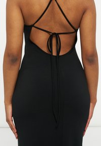 Γυναίκα που φοράει μαύρο φόρεμα χωρίς πλάτη με λεπτούς σταυρωτούς ιμάντες δεμένους με φιόγκο στη χαμηλή πλάτη, στέκεται μπροστά σε απλό φόντο.