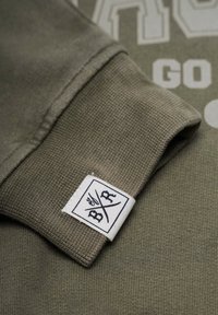 Lähikuva oliivinvihreän hupparin hihasta, jossa on valkoinen bränditagi, jossa on ristissä olevat kirjaimet "B" ja "R."