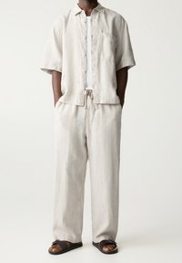 Lyhyet hihaiset pellavapaita ja siihen sopivat housut vaaleassa beigeessä, joissa on nappikiinnitys, rintatasku ja nyörivyötärö. Asusteena tummat sandaalit.