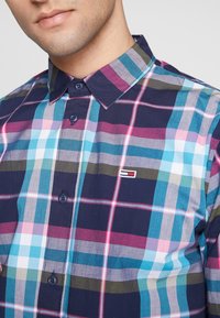 Chemise à manches courtes, à boutons, avec un motif à carreaux multicolores comprenant du bleu marine, du rose, du turquoise et du gris, dotée d'un petit logo de marque sur la poitrine.