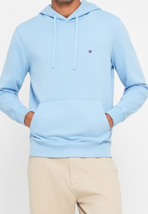 Mężczyzna w jasnoniebieskiej bluzie z kapturem z przednią kieszenią i małym logo Tommy Hilfiger, w połączeniu z beżowymi spodniami, ręce w kieszeniach.