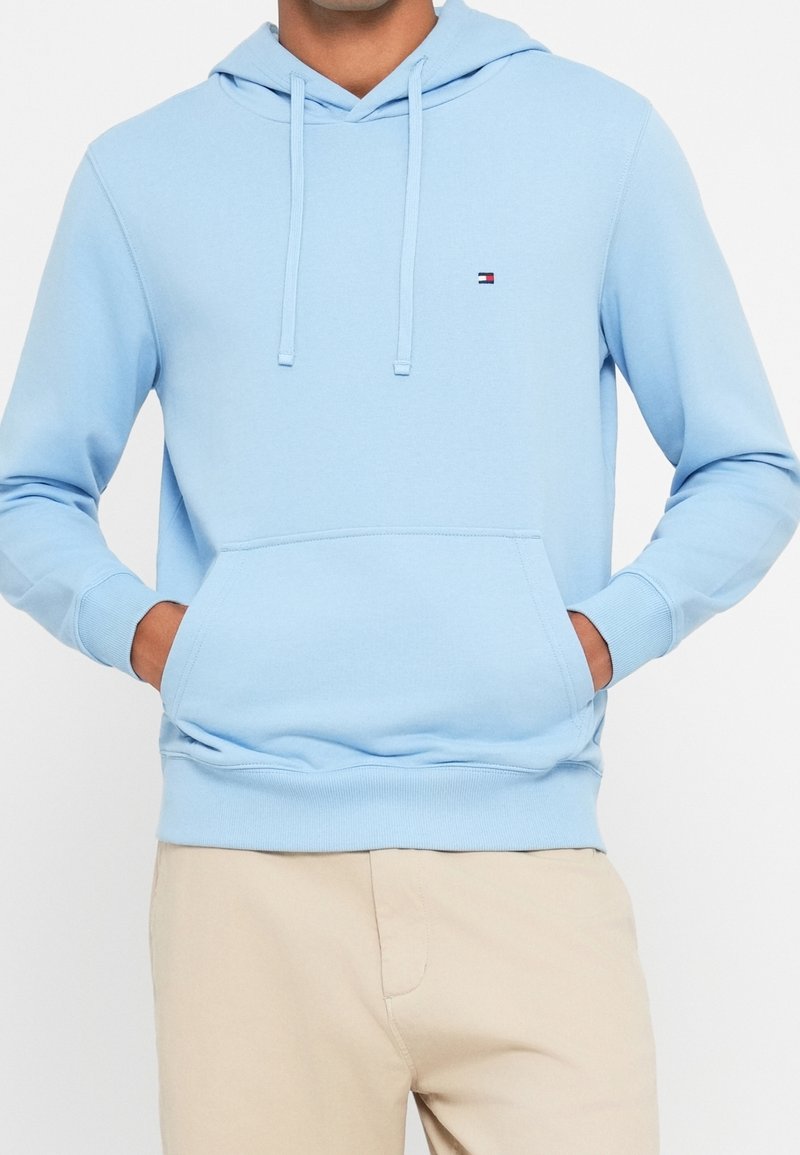 Homme portant un sweat à capuche bleu clair avec une poche avant et un petit logo Tommy Hilfiger, associé à un pantalon beige, mains dans les poches.