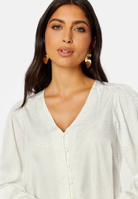 Blusa bianca con scollatura a V, motivo testurizzato e bottoni sul davanti. Maniche lunghe a sbuffo, tessuto morbido e leggera lucentezza.