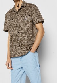 Košile s leopardím vzorem, krátký rukáv, v hnědé a černé barvě, s dvěma předními kapsami, uzávěrem na knoflíky a volným střihem. V kombinaci se světle modrými džíny.