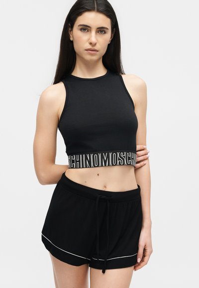 Žena ve černém bezrukávovém crop topu s bílým logem a černých šortkách s bílým lemováním, stojící před jednoduchým světlým pozadím.
