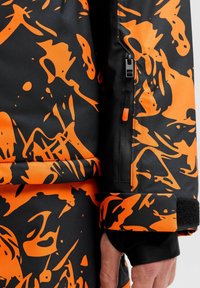Chaqueta con un patrón negro y naranja, que presenta una textura suave, un bolsillo con cremallera y acentos en el puño de Velcro. Diseño abstracto audaz con colores vibrantes.
