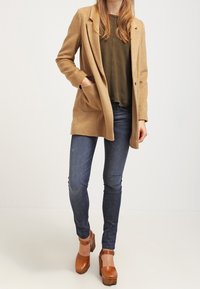 Abrigo de lana beige con solapas y bolsillos laterales, blusa verde oliva, jeans ajustados oscuros y tacones de plataforma marrones. Textura suave, corte entallado.