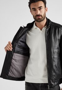 Zwarte leren jas met geribbeld kraag, rits sluiting en gestikte details. Binnenkant heeft een grijze voering en zakken. Draag over een witte polo.
