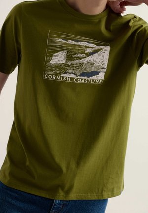 Person iført en olivengrøn skjorte med en hvid og sort grafik af et kystlandskab og teksten "Cornish Coastline" foran.