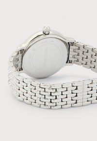 Ρολόι Swarovski από ατσάλινο ανοξείδωτο ασημί με συνδεδεμένο βραχιόλι και στρογγυλό καντράν που δείχνει χαραγμένο το λογότυπο και τις προδιαγραφές στο πίσω μέρος.