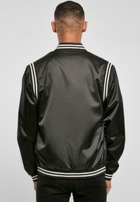 Čierna bomber bunda vyrobená zo saténu s bielymi pruhmi na rukávoch a golieri. Má priliehavý pás a dlhé rukávy.