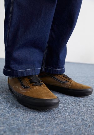 Sneakers marroni e nere con lacci indossate con jeans blu scuro a gamba larga su un pavimento con moquette blu.