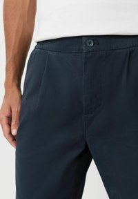 Námorně modré kalhoty vyrobené z lehkého materiálu, s knoflíkovým zapínáním, záhyby a hladkou texturou. Detailní pohled s umístěním ruky.