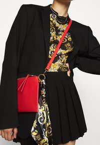 Czerwony torebka na ramię o eleganckim designie, z złotymi akcesoriami i dekoracyjną chustą. Tło jest jednolicie białe.
