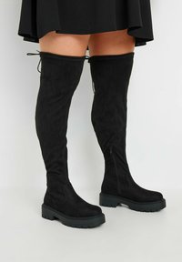 Svarta knähöga stövlar i mjukt mocka, med en tjock svart gummisula, sidodragkedja och justerbara snören upptill.