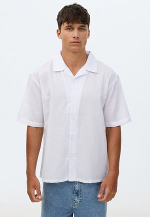 Joven con camisa blanca de manga corta abotonada y vaqueros azul claro, de pie frente a un fondo claro y liso.