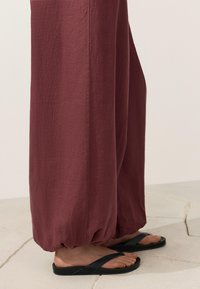 Personne portant un pantalon large bordeaux avec des poignets élastiques et des sandales plates noires, debout sur un sol carrelé clair.