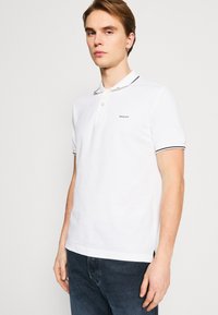 Witte polo met een knoopsluiting met twee knopen, zwarte accenten op de kraag en mouwen, voorzien van een klein geborduurd logo op de borst.