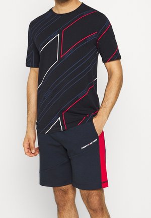 Man draagt een zwart T-shirt met geometrisch patroon en marineblauwe korte broek met rode zijpanelen en Tommy Hilfiger-logo.