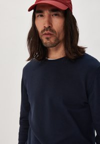 Tamsiai mėlynas ilgomis rankovėmis megztinis su apvaliu iškirpimu, pagamintas iš švelnios medžiagos. Dėvimas su raudona kepure, kuri turi lygų paviršių ir prigludusią formą.