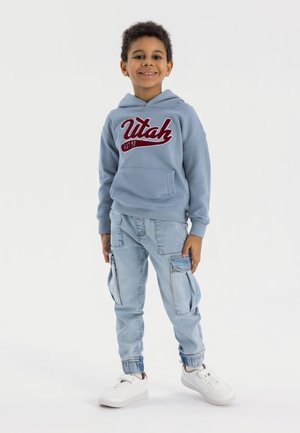 Felpa blu chiaro con logo "Utah" in bordeaux, abbinata a pantaloni cargo in denim chiaro con molteplici tasche e polsini elasticizzati, sneakers bianche.