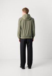 Chaqueta verde oliva con capucha, puños y cintura acanalados. Pantalones negros de corte relajado. Fondo liso sin detalles adicionales.