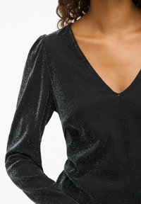 Svart långärmad topp med V-ringning. Tillverkad av glittrande tyg med subtil textur och glänsande detaljer. Har puffade ärmar för struktur.