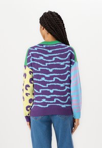 Pull tricoté avec un mélange de motifs en violet, vert, jaune, bleu et rose. Col rond, dos allongé, manches au design contrasté.