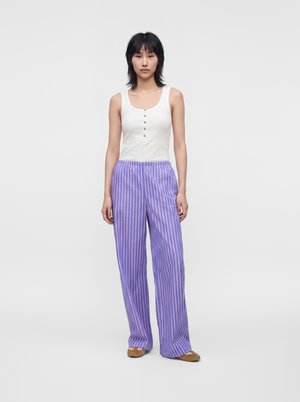 Femme debout face à l'avant, porte un haut blanc sans manches à boutons et un pantalon large à rayures violettes avec des chaussures marron sur un fond uni.
