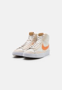 Höga sneakers i beige läder med en orange Nike-swoosh. Har vita snören och en strukturerad vit gummisula.