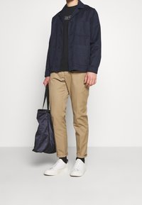 Muž v námořnickém saku, černé košili, béžových kalhotách a bílých teniskách, držící námořnickou tašku na pozadí čistě bílé.