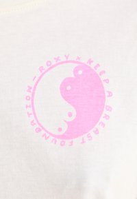 Lysecreme t-shirt med et cirkulært rosa logo med yin-yang symbol og teksten "ROXY x KEEP A BREAST FOUNDATION" på fronten.