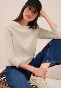 Frau mit langen braunen Haaren, die eine schwarze Mütze, ein elfenbeinfarbenes, geripptes Langarmoberteil, blaue Jeans und hellbeige Sneaker trägt, sitzt auf einer Holzbank.