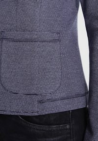 Mörkblå och vit randig jacka med rundad ficka, skräddarsydd passform och texturerat tyg. Närbild av tyget och sömnadsdetaljer.