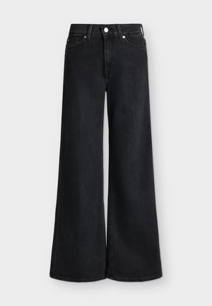 Femme debout portant une veste en cuir grise, un pantalon large noir, des tops noirs et blancs superposés, tenant une pochette en cuir de serpent.