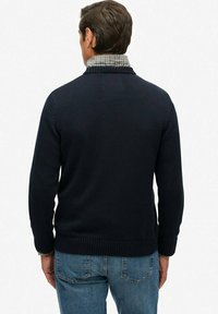 Pull en tricot bleu marine à col haut, avec une doublure intérieure en tissu à carreaux. Porté avec un jean en denim bleu clair. Tissu texturé.
