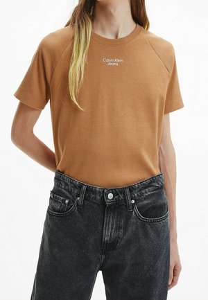 Osoba v hnědém tričku s krátkým rukávem s nápisem "Calvin Klein Jeans" a černých denimových kalhotách s vysokým pasem na jednoduchém bílém pozadí.