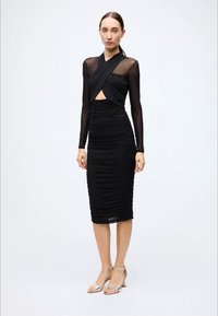Black ruched dress with long sheer sleeves, a cross-over bodice, and a mid-length skirt. Features cut-out detail at the waist.