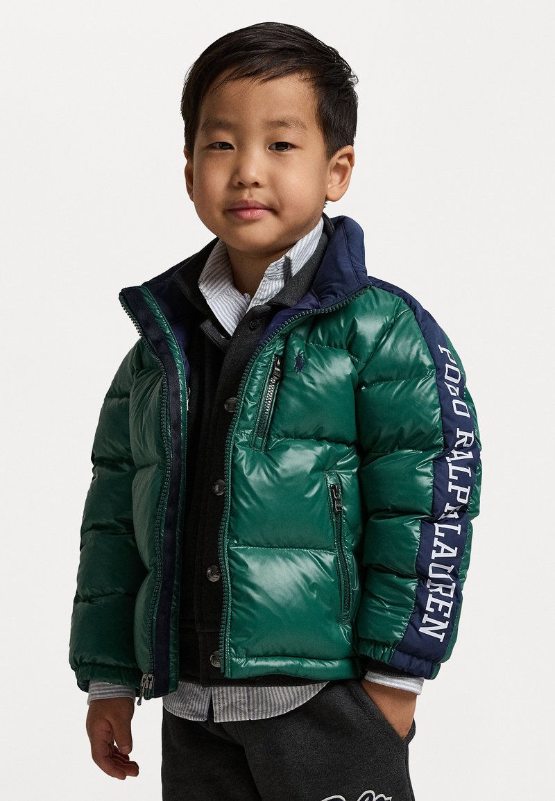 Groene pufferjack met marineblauwe accenten, rits- en knoopsluiting aan de voorkant, logodetail op de mouw, gedragen over een grijs sweater en gestreept shirt.