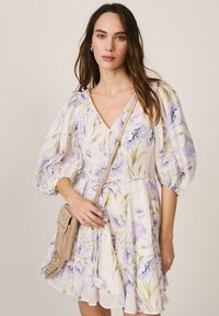 Mujer con un vestido blanco de flores, mangas abullonadas y un bolso cruzado beige con tachuelas, sobre un fondo neutro.