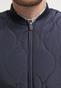 Chaleco bomber acolchado en color navy con cuello acanalado, cremallera expuesta y sutiles patrones ondulados en la tela para añadir textura.