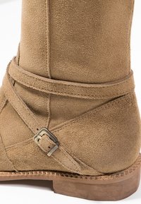 Suede ankle boot in light brown with a cross-strap design. Features a metal buckle accent and a textured surface on the shaft.
