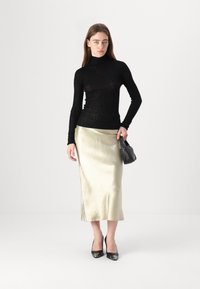 Maglietta nera a coste con maniche lunghe abbinata a una gonna midi in raso di colore oro metallizzato. Modella tiene una piccola borsa nera a tracolla, indossando tacchi neri.