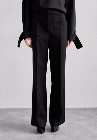 Pantaloni neri a gamba larga realizzati in un tessuto liscio, con grandi tasche posteriori e vita alta. Indossati con una maglia nera ampia e scarpe con tacco alto.