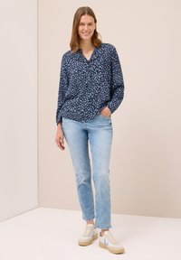 Blusa navy con un diseño estampado y escote en V, combinada con jeans de color azul claro y zapatillas blancas con acentos azules.