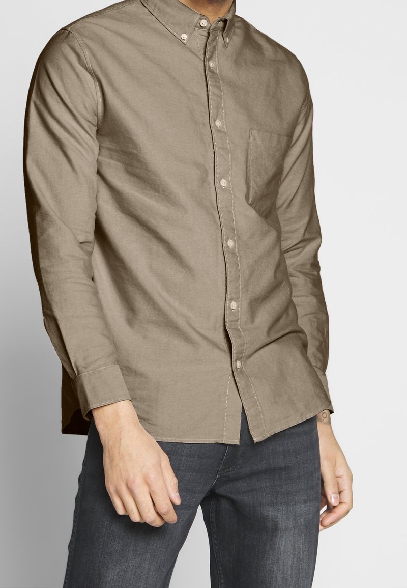 Homme portant une chemise beige à manches longues boutonnée et un jean bleu foncé, debout devant un fond gris clair uni.