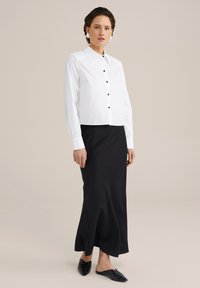 Camicia bianca a maniche corte con bottoni neri, abbinata a una lunga gonna nera liscia e a scarpe slip-on nere. Design minimalista.