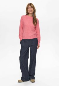 Roza pletena jopica z dolgimi rokavi, okroglih izrezom in rebrastimi manšetami, v kombinaciji z modrimi širokimi denim hlačami in bež obutvijo.