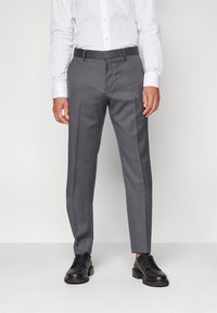Pantalons gris ajustés avec plis, fabriqués en tissu lisse, présentant une coupe droite et des passants de ceinture, associés à des chaussures en cuir noires.
